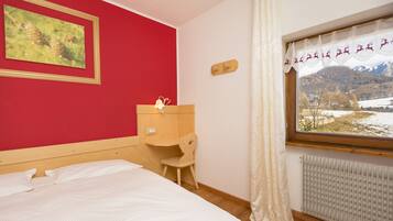A bedroom with a bed, a wooden nightstand, a chair, a window with a view of a snowy landscape, and a red wall with a framed picture.