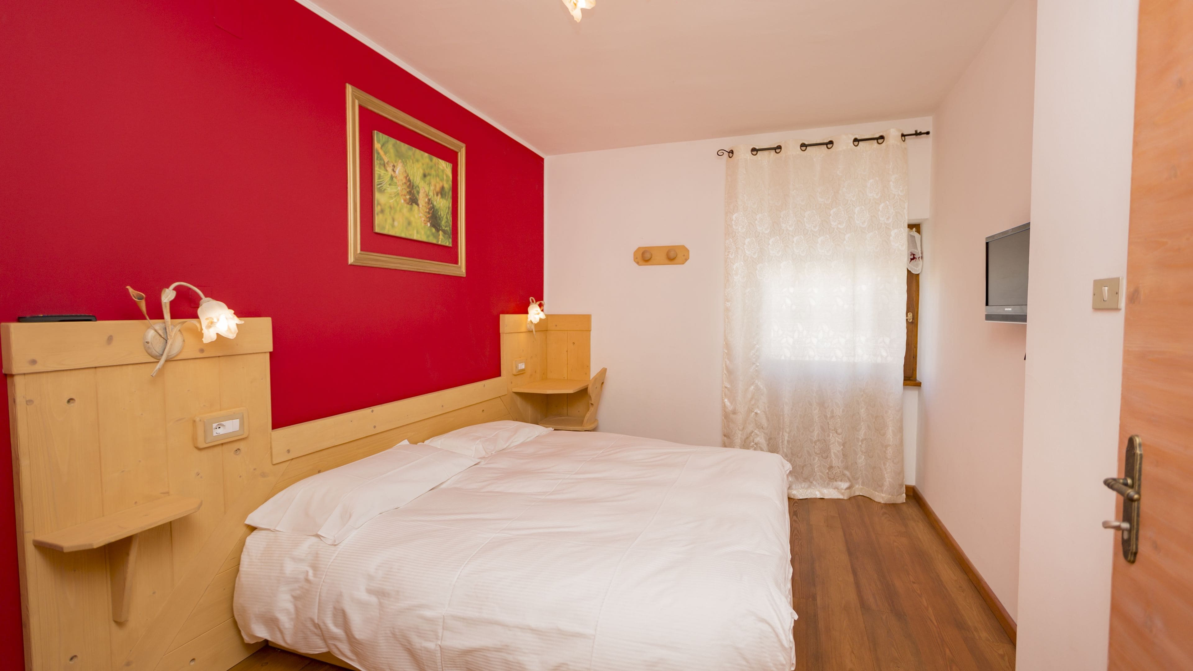 Un dormitorio con cabecera de madera, una cama individual, pared roja, un cuadro enmarcado, una mesita de noche con lámpara y una ventana con cortina.