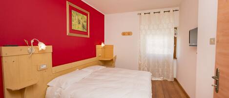 A bedroom with a wooden headboard, a single bed, a red wall, a framed picture, a bedside table with a lamp, and a window with a curtain.