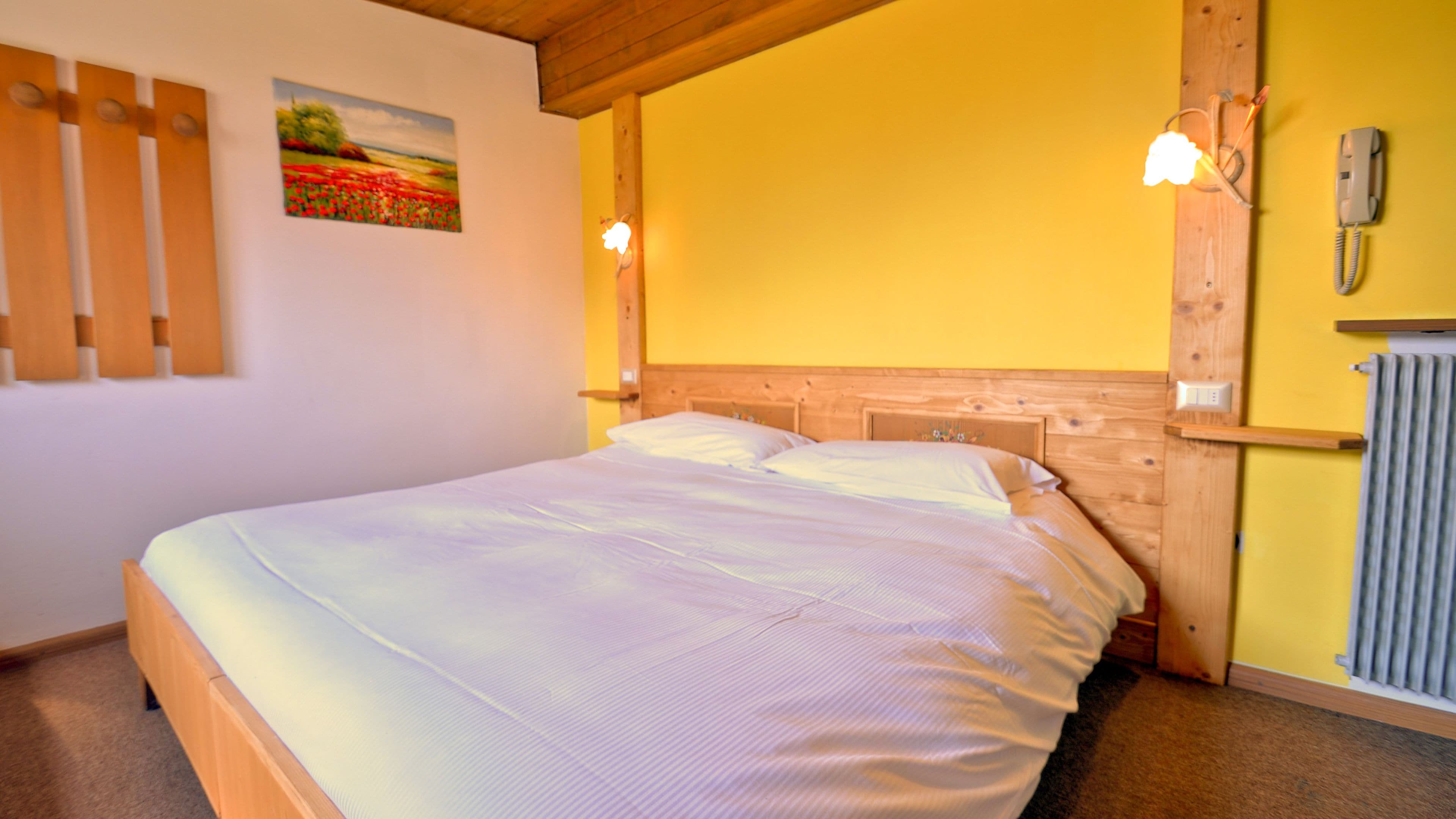 A bedroom with a wooden headboard, a bed with white bedding, a painting on the wall, and a telephone on the wall.