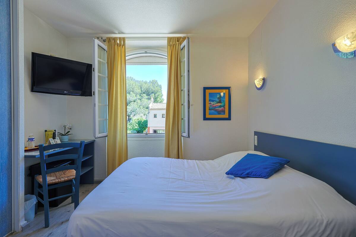 A hotel room with a bed, a desk with a chair, a TV mounted on the wall, and a window with curtains.