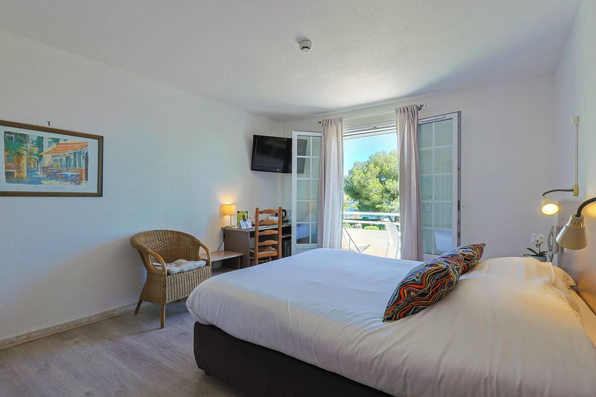 A bedroom with a bed, a chair, a desk, a TV, and a balcony with a view of trees.