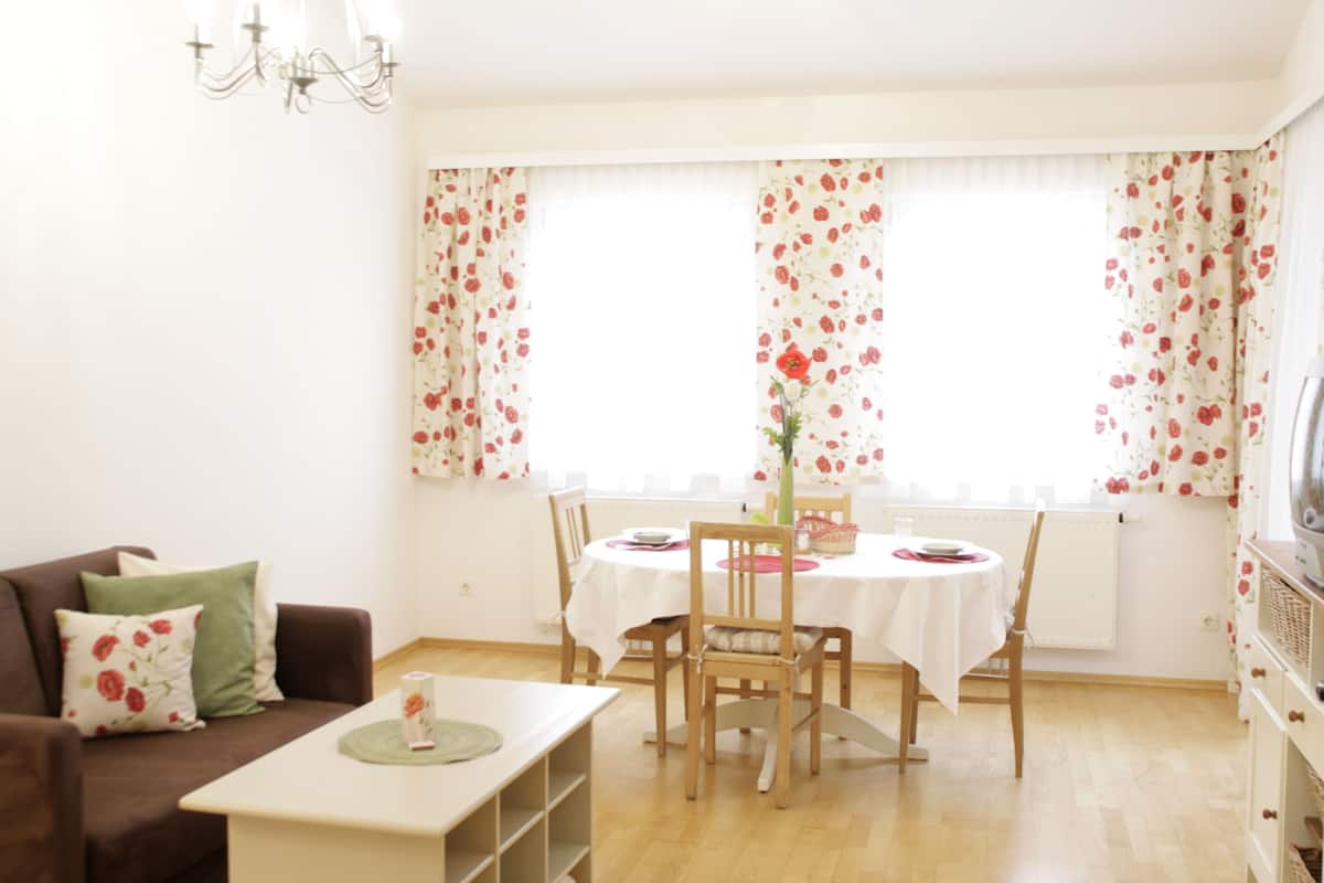 Uma sala de jantar com uma mesa posta para quatro pessoas, rodeada por cadeiras, um sofá e uma televisão.