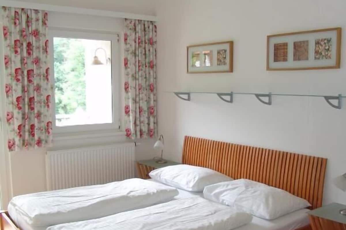 Uma cama de casal com cabeceira de madeira, uma janela com cortinas florais e duas imagens emolduradas na parede.