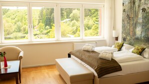 Un dormitorio con un ventanal grande, una cama con almohadas y una colcha, una silla y una mesa con un jarrón.