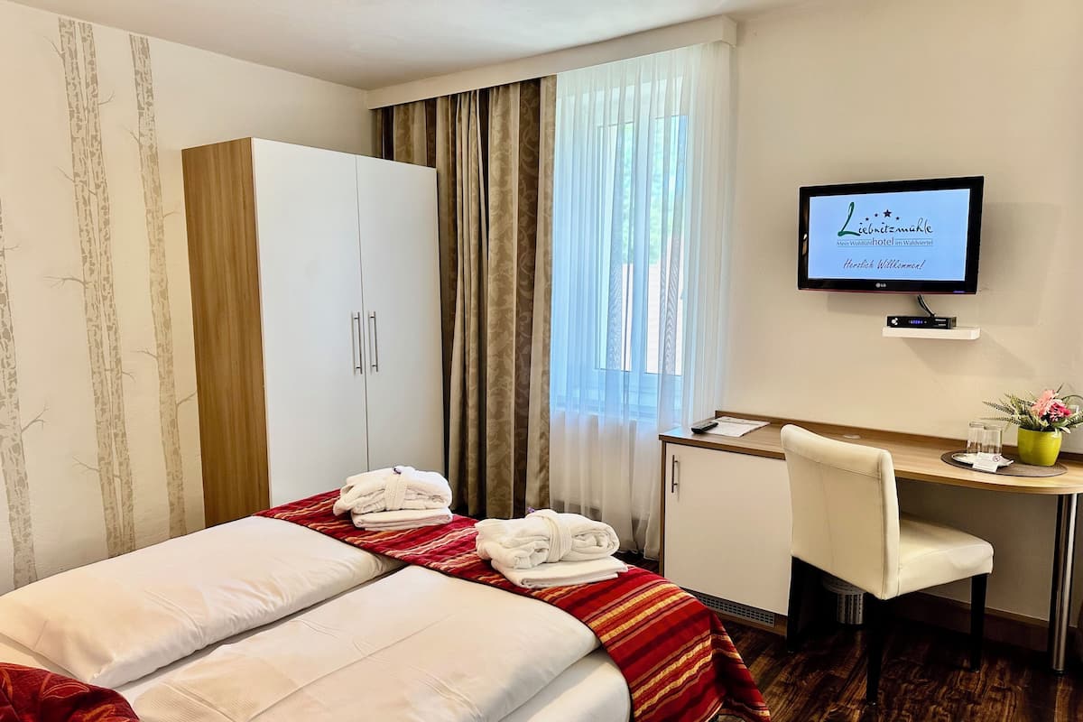 Quarto de hotel com uma cama, uma escrivaninha, uma cadeira e uma televisão exibindo uma mensagem de boas-vindas.
