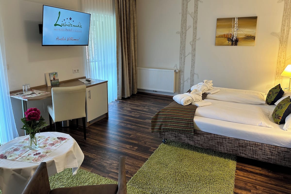 Quarto de hotel com cama, escrivaninha, cadeira, mesa com um vaso de flores e TV exibindo uma placa do hotel.