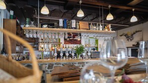 Zona de bar con estantes llenos de botellas y vasos, mobiliario de madera y lámparas colgantes.