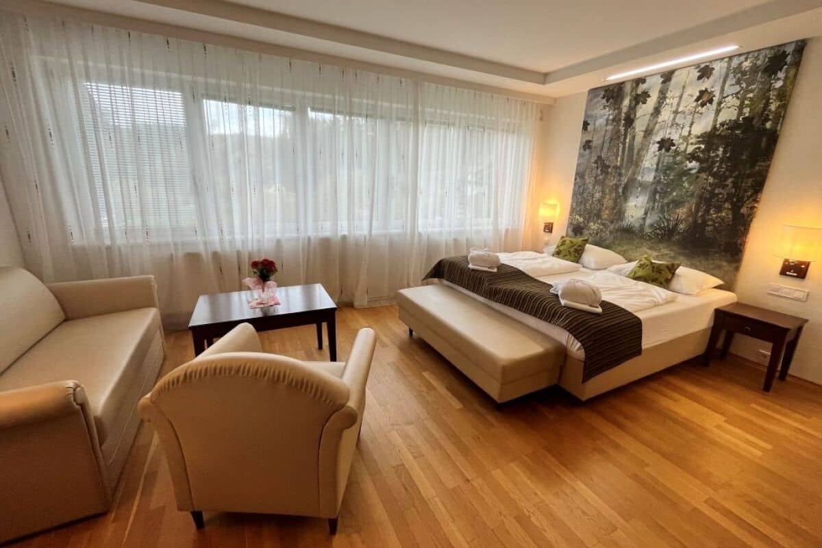 Quarto de hotel com uma cama, um sofá, uma cadeira, uma mesinha e um quadro grande com tema de floresta na parede.