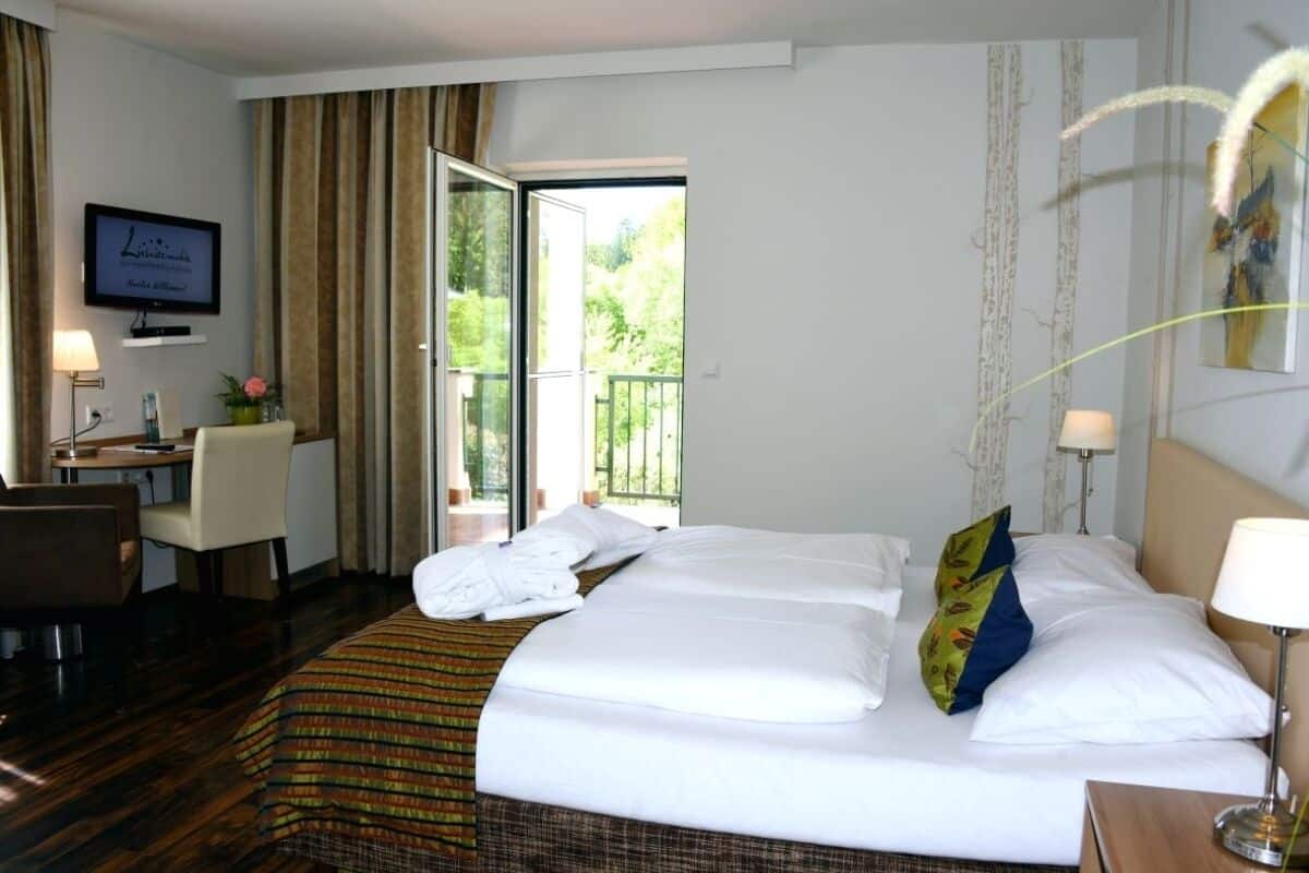 Quarto de hotel com cama, escrivaninha, cadeira, televisão e varanda com cortinas.