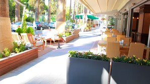 An outdoor seating area with wicker furniture and planters, a covered walkway, and a view of parked cars and trees. - Karaca Otel (Izmir)