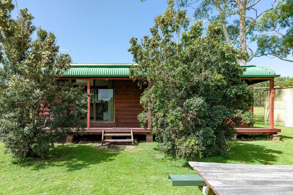 A rustic cabin with a green roof, surrounded by lush greenery and a clear blue sky.