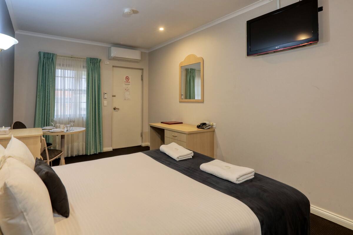 Quarto de hotel com cama, escrivaninha, cadeira, mesa com copos e um livro, espelho e televisão.