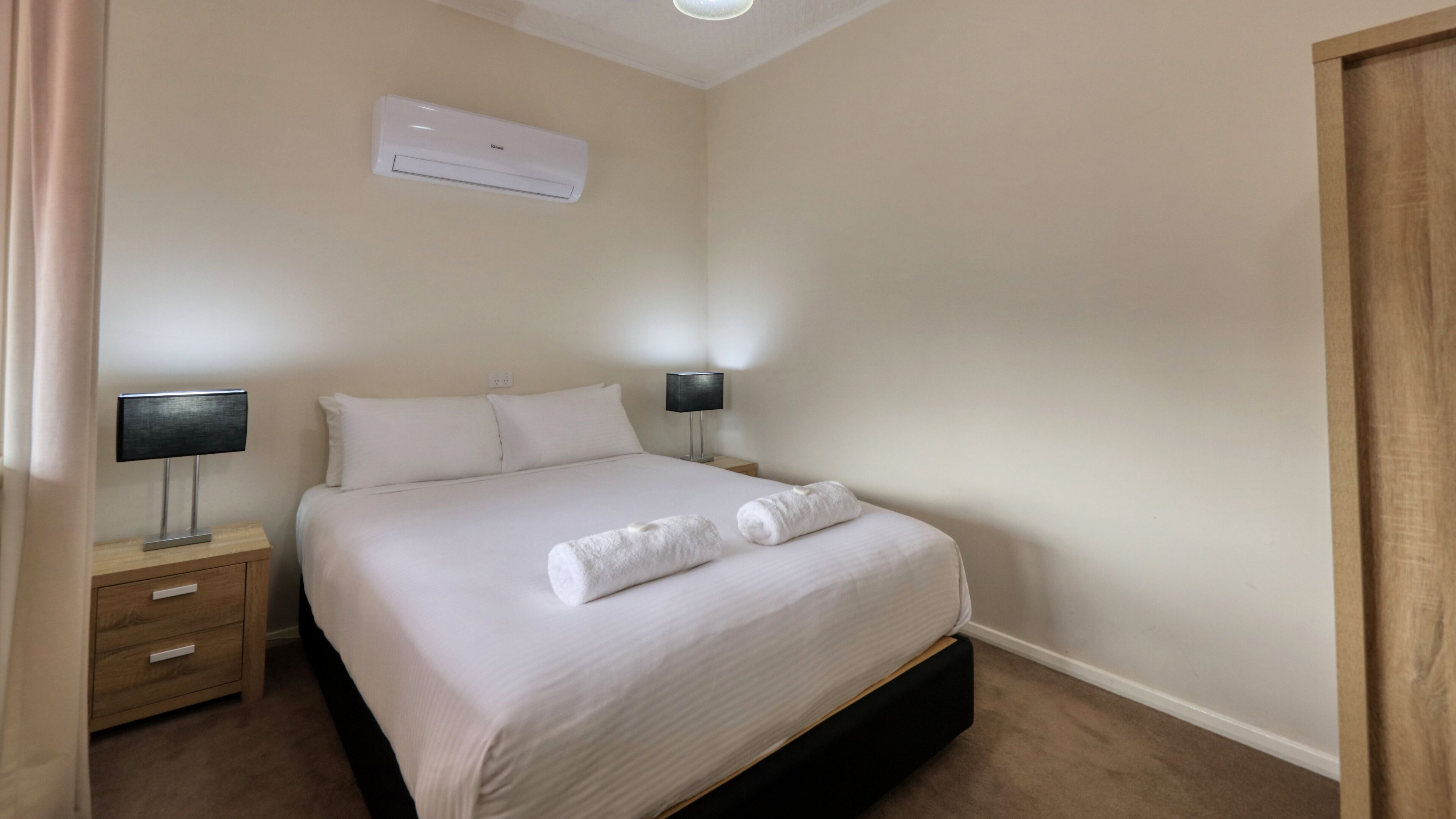 Ein ordentlich bezogenes Bett mit weißen Leinen und zwei aufgerollten Handtüchern, ein Nachttisch mit einer Lampe und eine Wand-Klimaanlage.