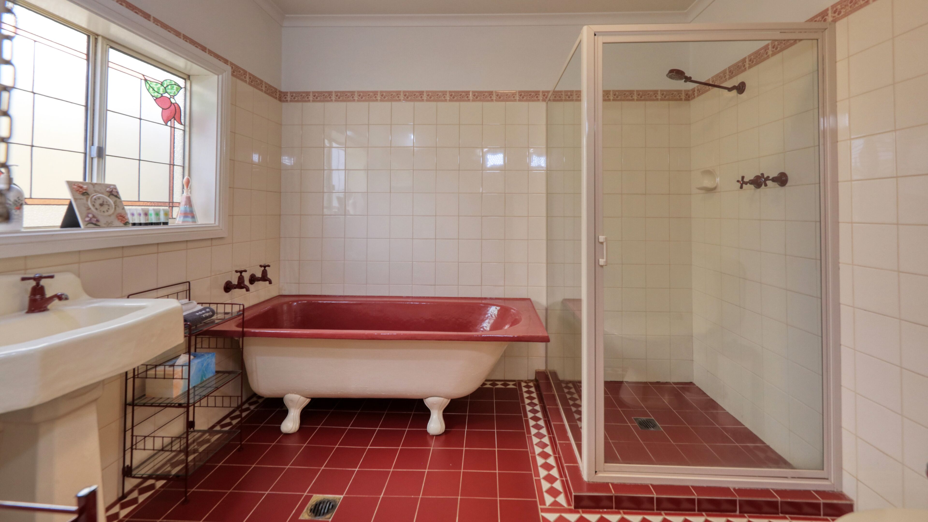 Une salle de bain avec un sol carrelé de carreaux rouges, une baignoire blanche, un espace douche et une fenêtre ornée de motifs floraux.