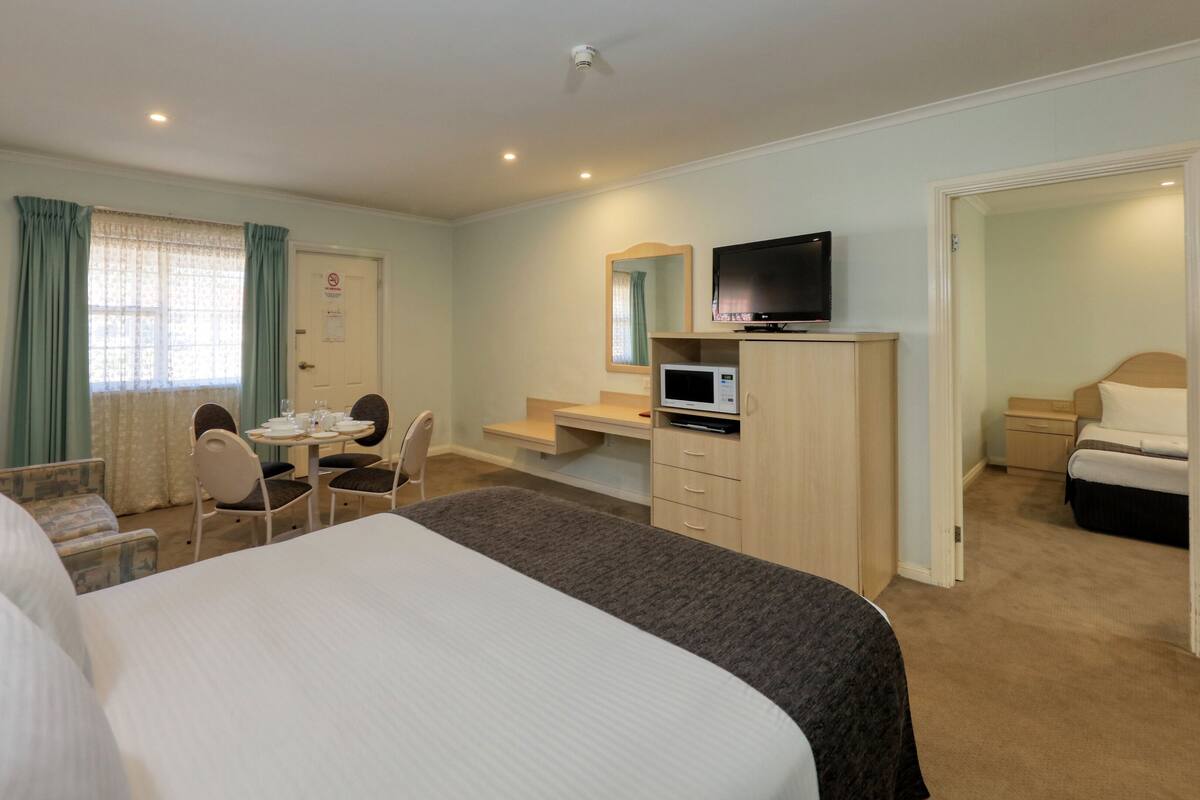 Quarto de hotel com uma cama, uma escrivaninha com computador, televisão, cadeira e uma mesinha com copos.