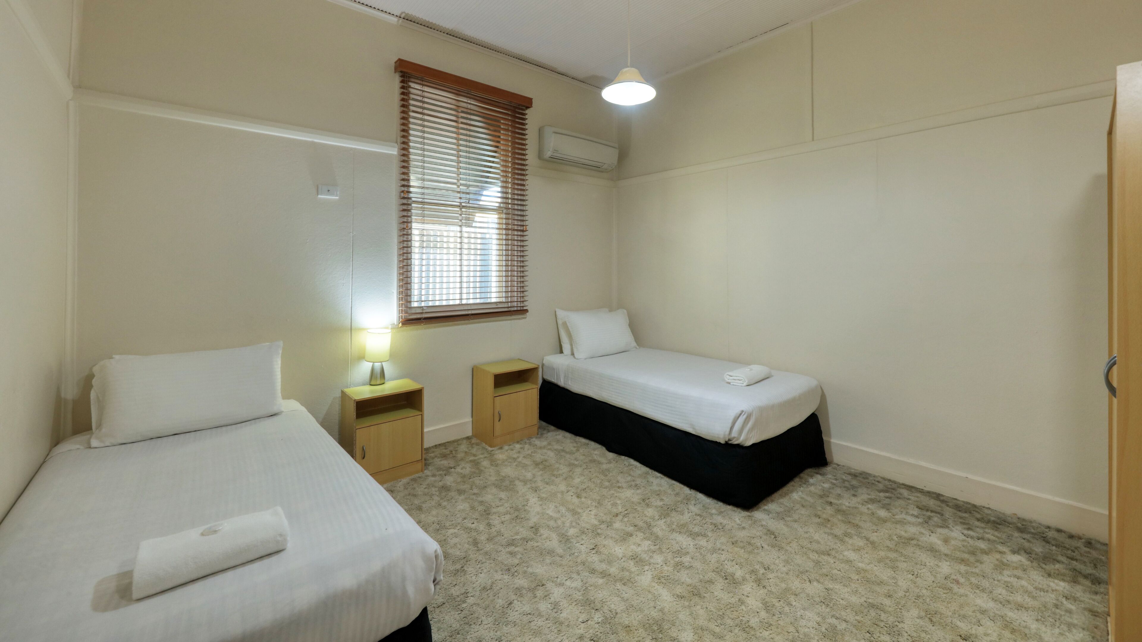 Une chambre avec deux lits simples, une petite table de chevet en bois avec une lampe, une fenêtre avec des stores et un appareil de climatisation fixé au mur.