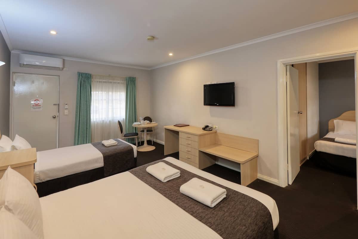 Quarto de hotel com duas camas, uma escrivaninha, uma cadeira, uma televisão e uma porta que dá para outro quarto.