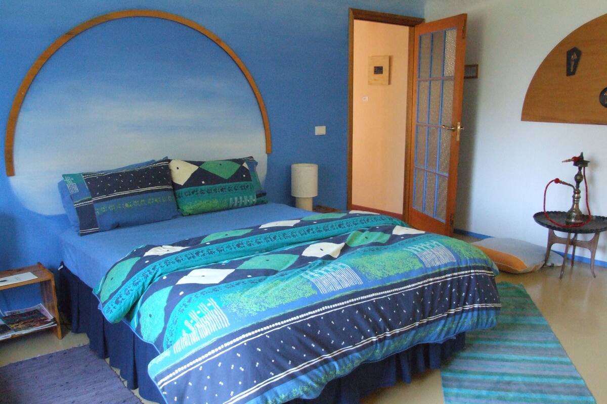 Un dormitorio con una cama, una mesita de noche, una lámpara, una puerta y una pequeña mesa con una cachimba.