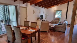 Amplia sala de estar con una mesa de comedor de madera, sillas de mimbre, un sofá color beige y un televisor sobre un soporte.