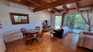 Una sala de estar acogedora con techo de madera, una mesa de comedor con sillas, un sofá, un televisor y un ventanal con cortinas.