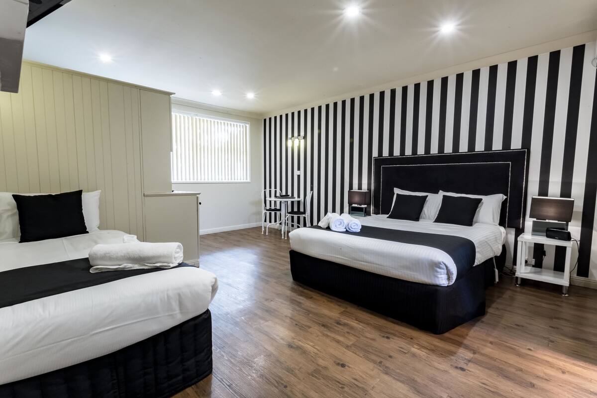 A hotel room with two beds, a large window, a striped wall, and a small table with a lamp.