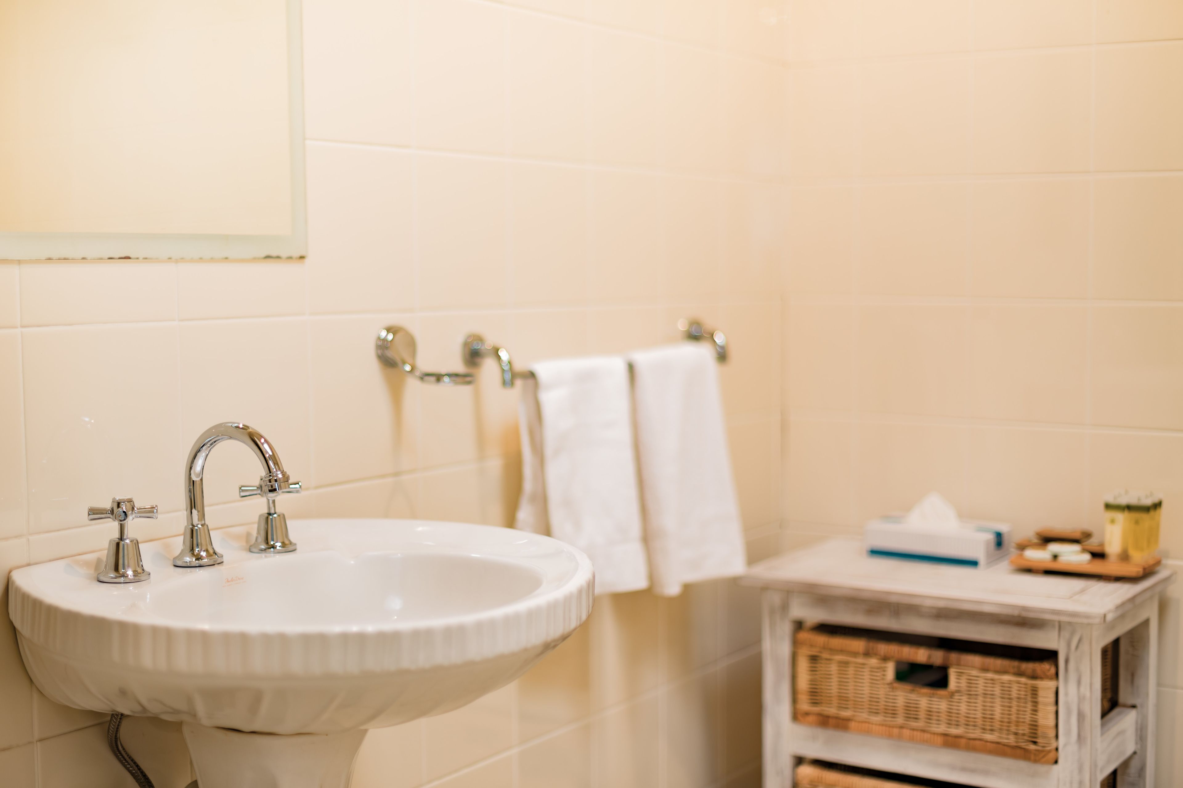 A bathroom with a white sink, two faucets, a mirror, and a towel rack with a white towel. There is a small wooden side table with a tissue box and other toiletries.