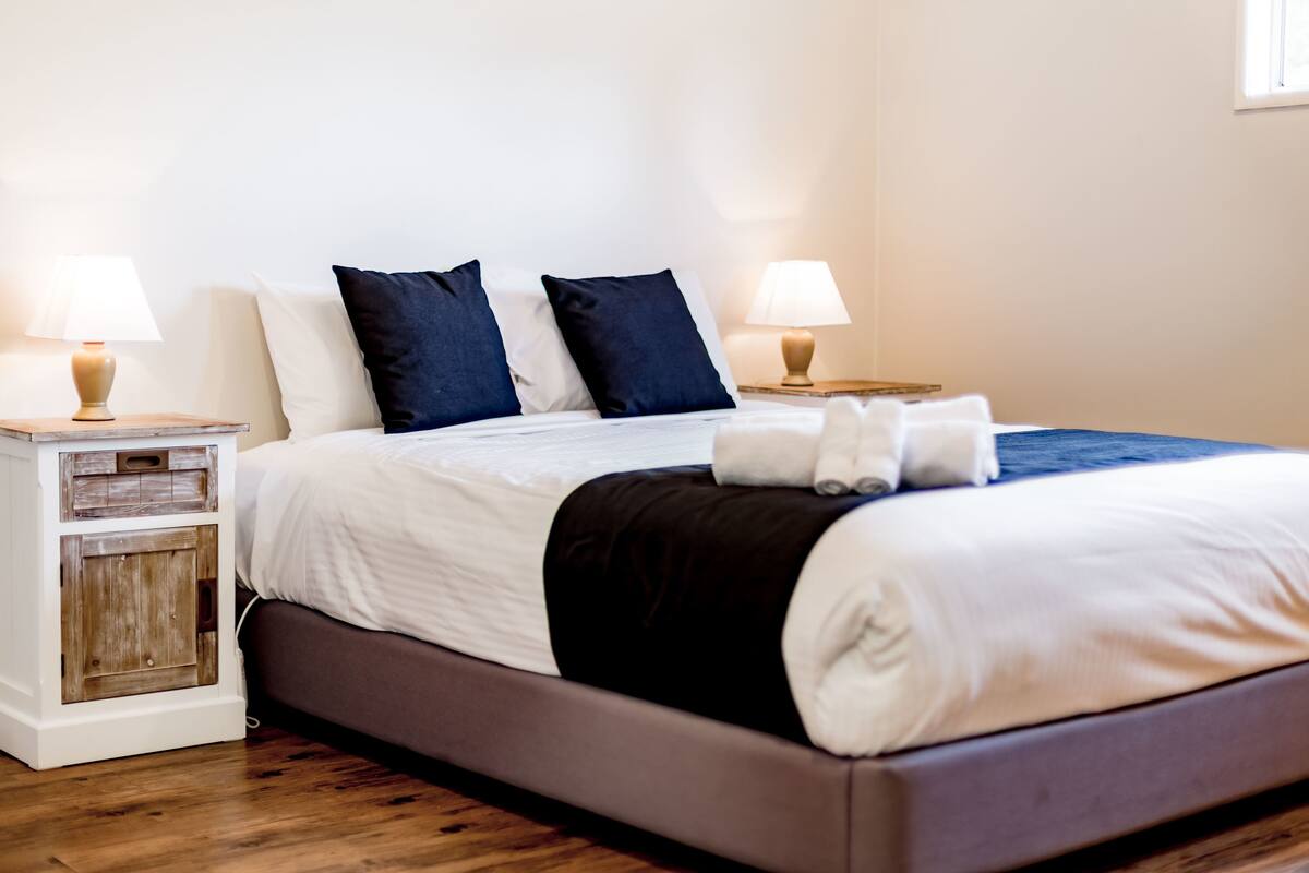 A neatly made bed with white and dark blue bedding, two bedside tables with lamps, and a wooden nightstand with drawers.