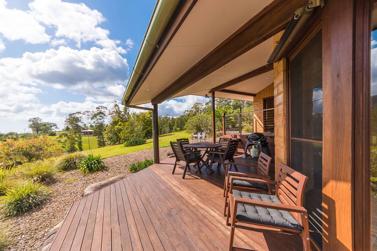 A wooden deck with outdoor furniture, a covered patio area, and a view of a landscaped garden and hills in the background.