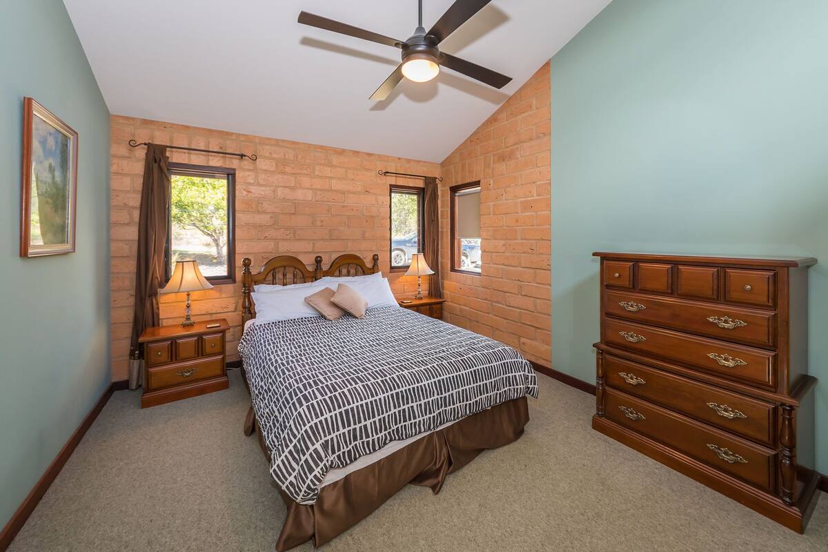 A bedroom with a bed, two nightstands, a dresser, a ceiling fan, and brick walls.