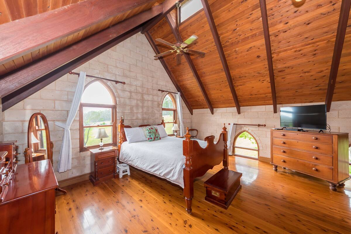 A bedroom with a wooden ceiling, a bed, a dresser, a television, and two windows with curtains.