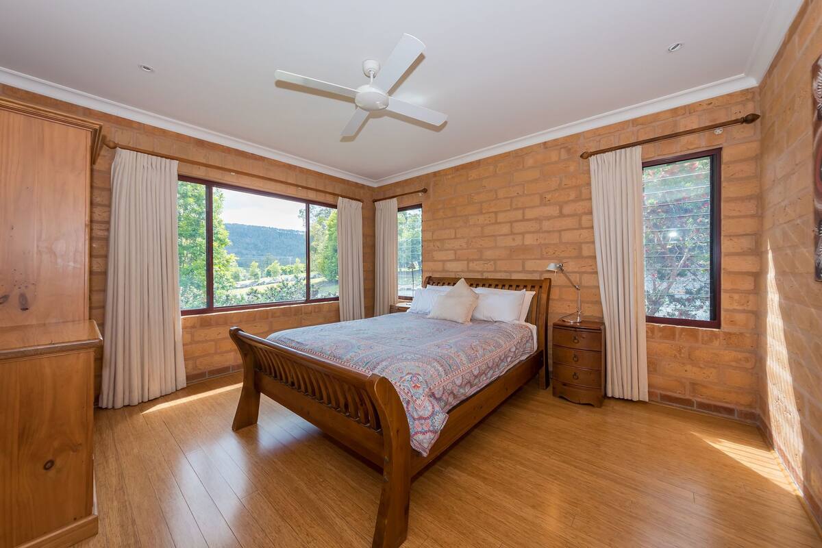 A bedroom with a wooden bed, a nightstand, and a ceiling fan. There are large windows with curtains, and a brick wall.