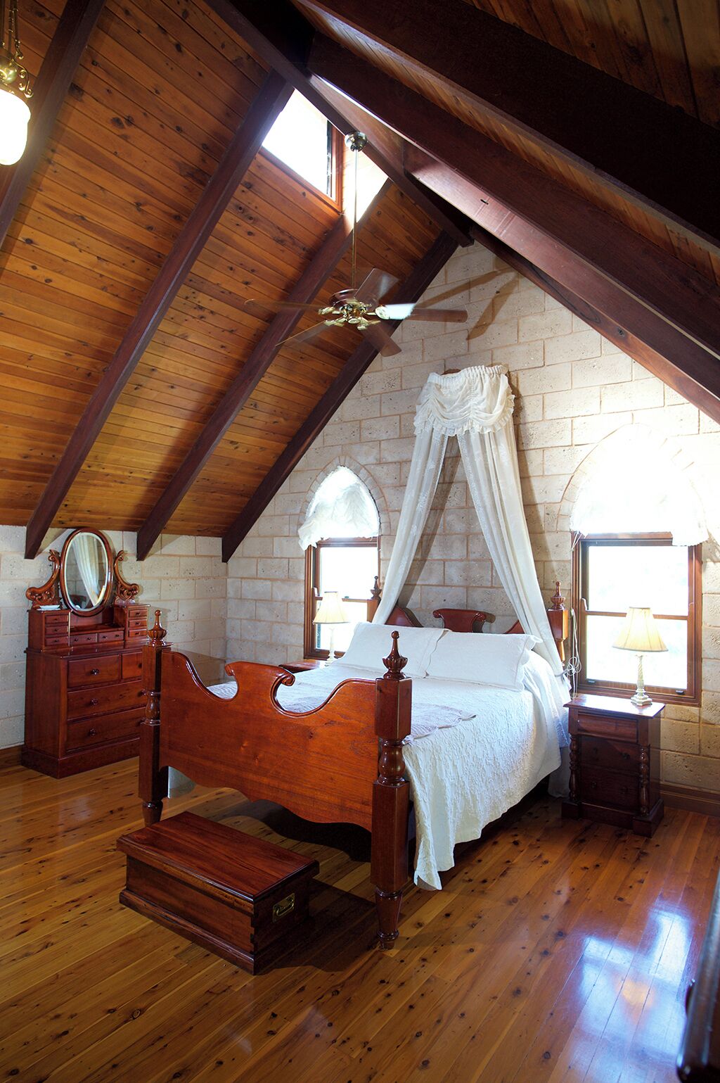 A bedroom with a wooden ceiling, a four-poster bed, a wooden dresser, a mirror, and a window with curtains.