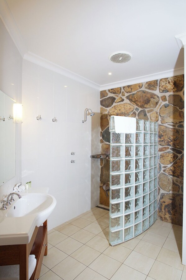 Un baño con un lavamanos blanco, dos espejos, ducha con mampara de vidrio y pared de piedra.