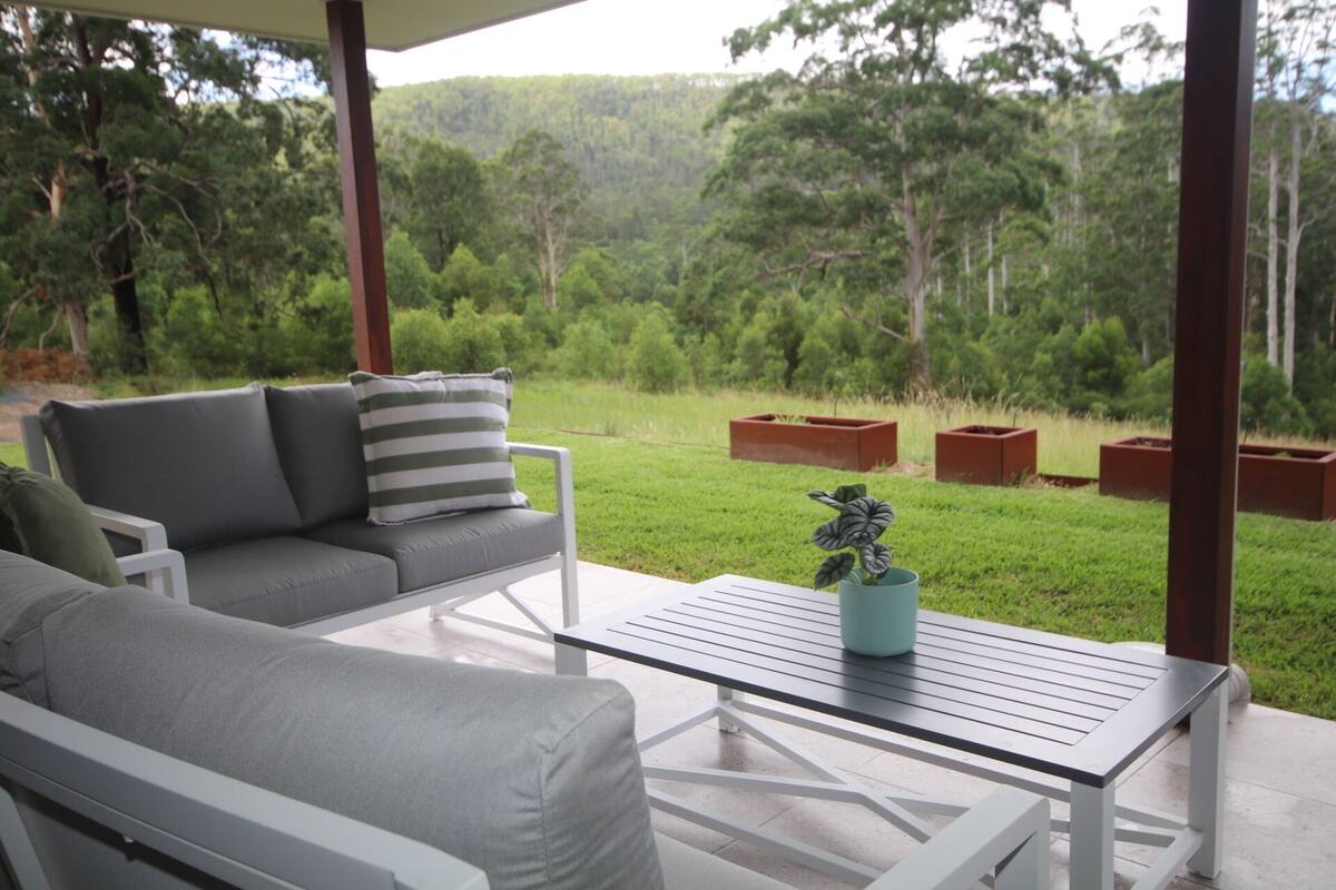 A covered outdoor seating area with a sofa and a table, surrounded by planters and a view of a grassy landscape with trees.