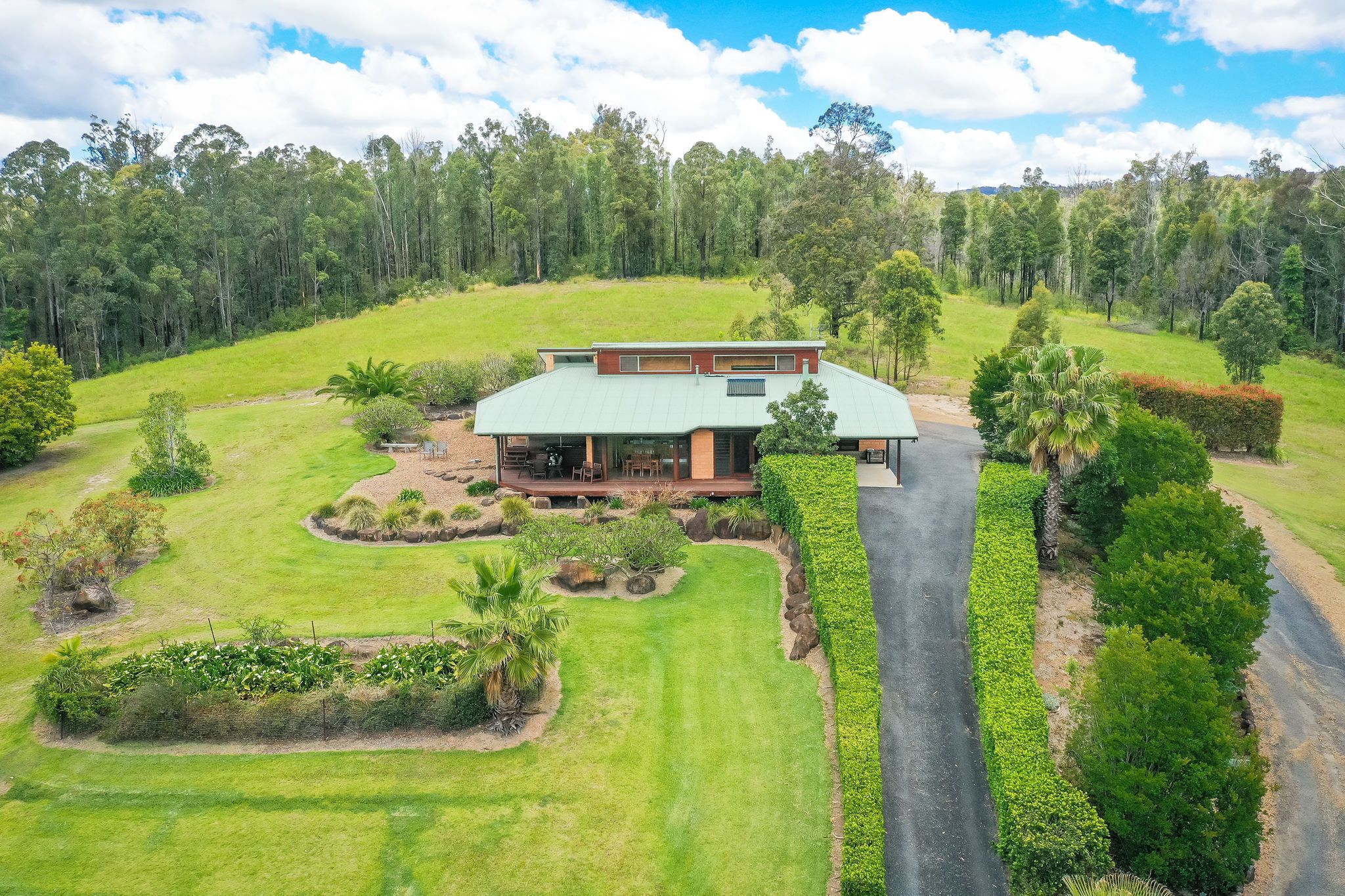 A house with a green roof, surrounded by a well-maintained garden and a forested hill in the background.