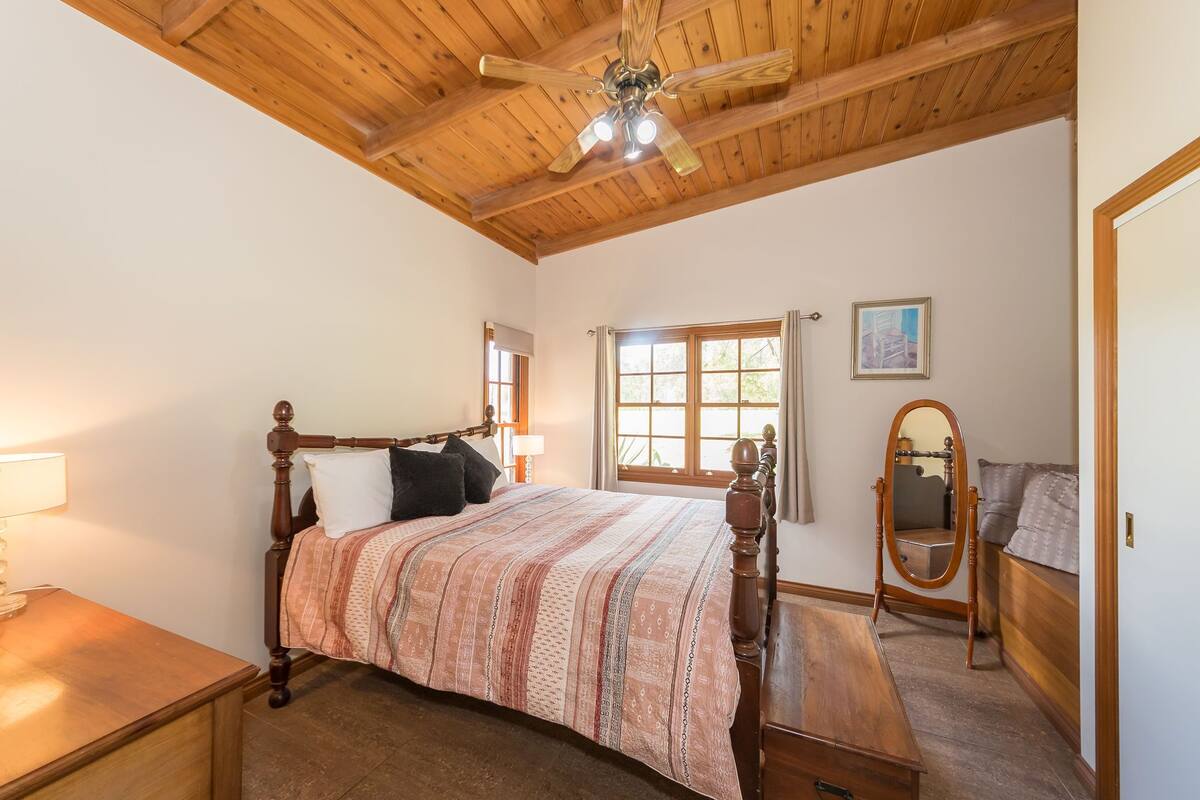 A bedroom with a wooden ceiling, a bed with a patterned bedspread, a nightstand, a dresser, a mirror, and a window with curtains.