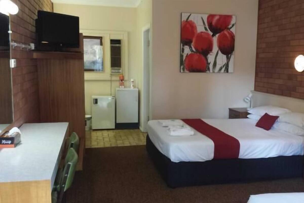 A hotel room with a bed, bedside table, and a small desk with a chair. There is a refrigerator and a microwave in the corner.