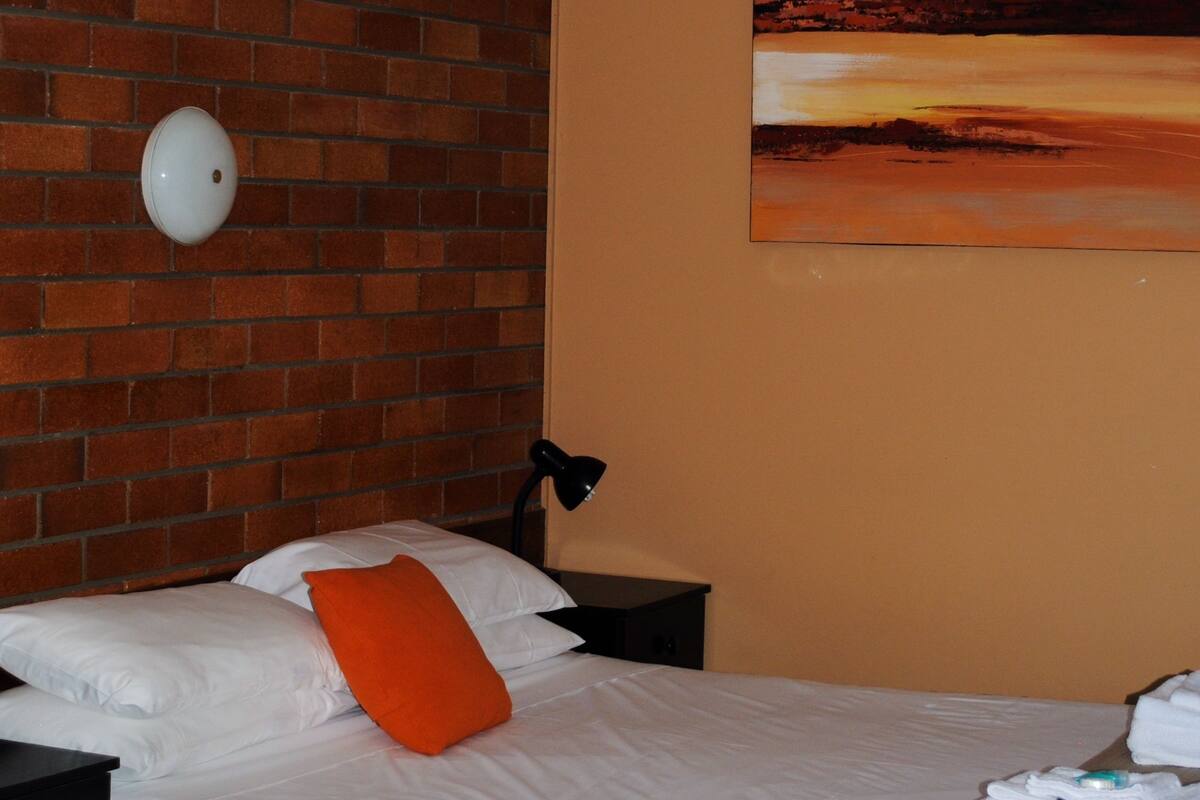 A hotel room with a brick wall, a bed with white bedding and an orange pillow, a nightstand with a lamp, and a painting of a sunset.