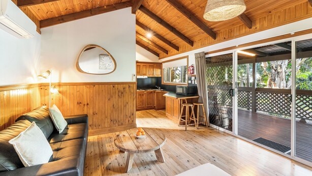 Un interior de madera con un sofá, una mesa redonda de madera y una zona de cocina con armarios de madera y un microondas.