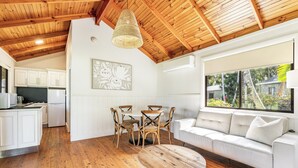 Una cocina amplia con armarios blancos, un comedor con mesa y sillas de madera, y un sofá blanco junto a un ventanal con vistas a zonas verdes.