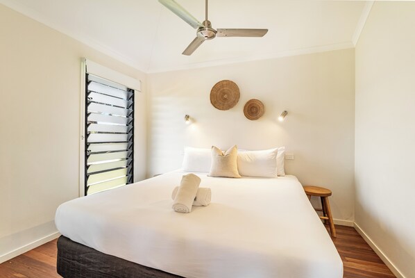 A neatly made bed with white linens and rolled towels, a ceiling fan, a wooden stool, and two wall-mounted light fixtures.