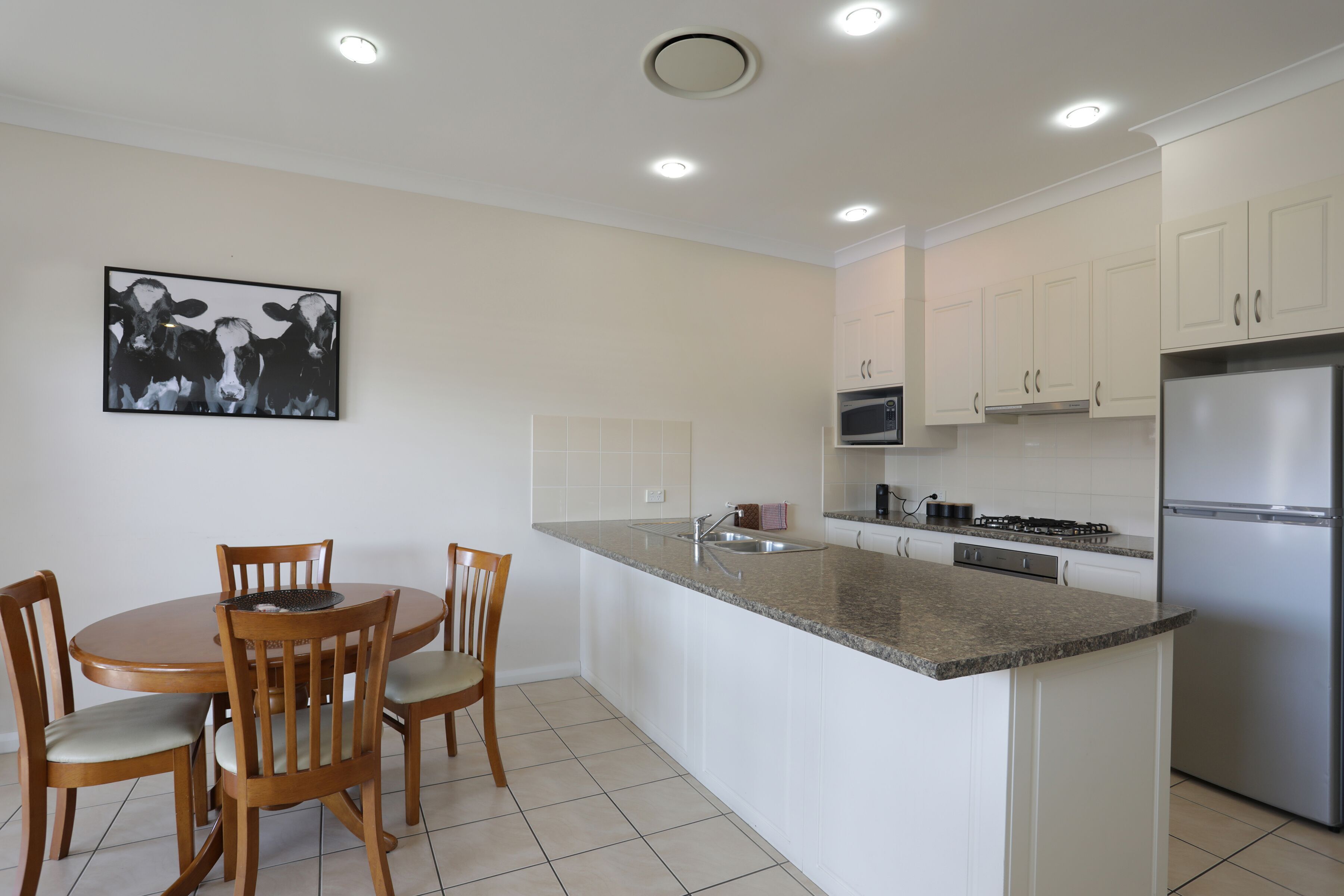 A modern kitchen with white cabinets, a stainless steel refrigerator, a microwave, and a dining area with wooden chairs and a table.