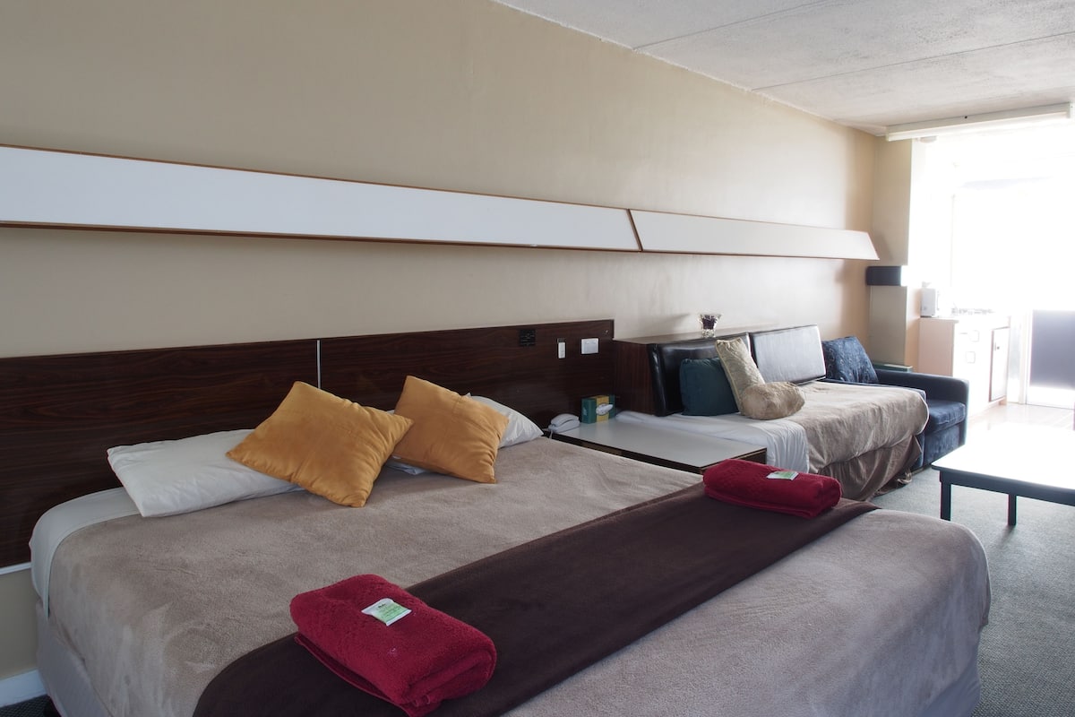 A hotel room with two beds, a sofa, a small table, and a view of the outdoors.