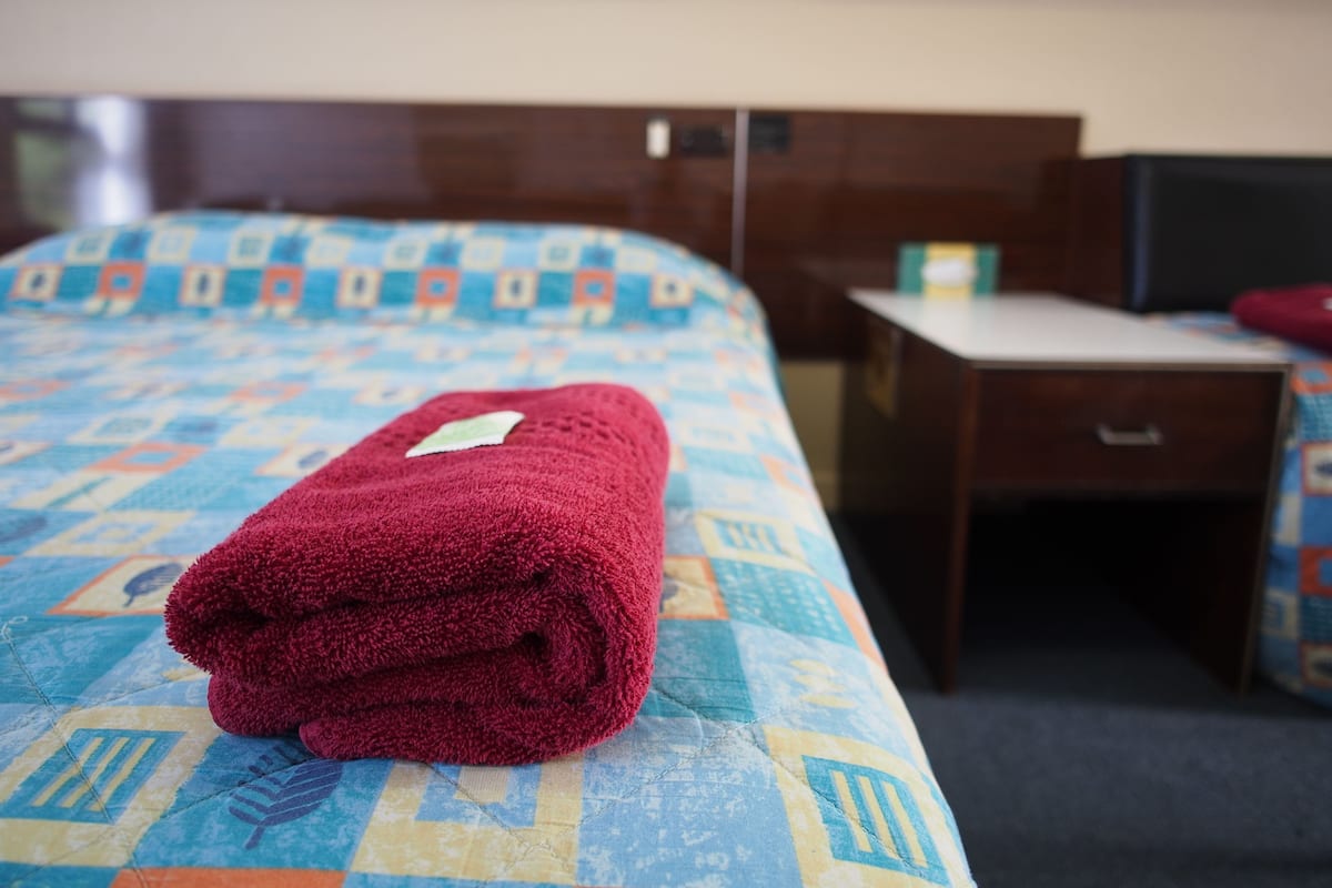 A neatly made bed with a red towel, a patterned bedspread, a small bedside table, and a nightstand.