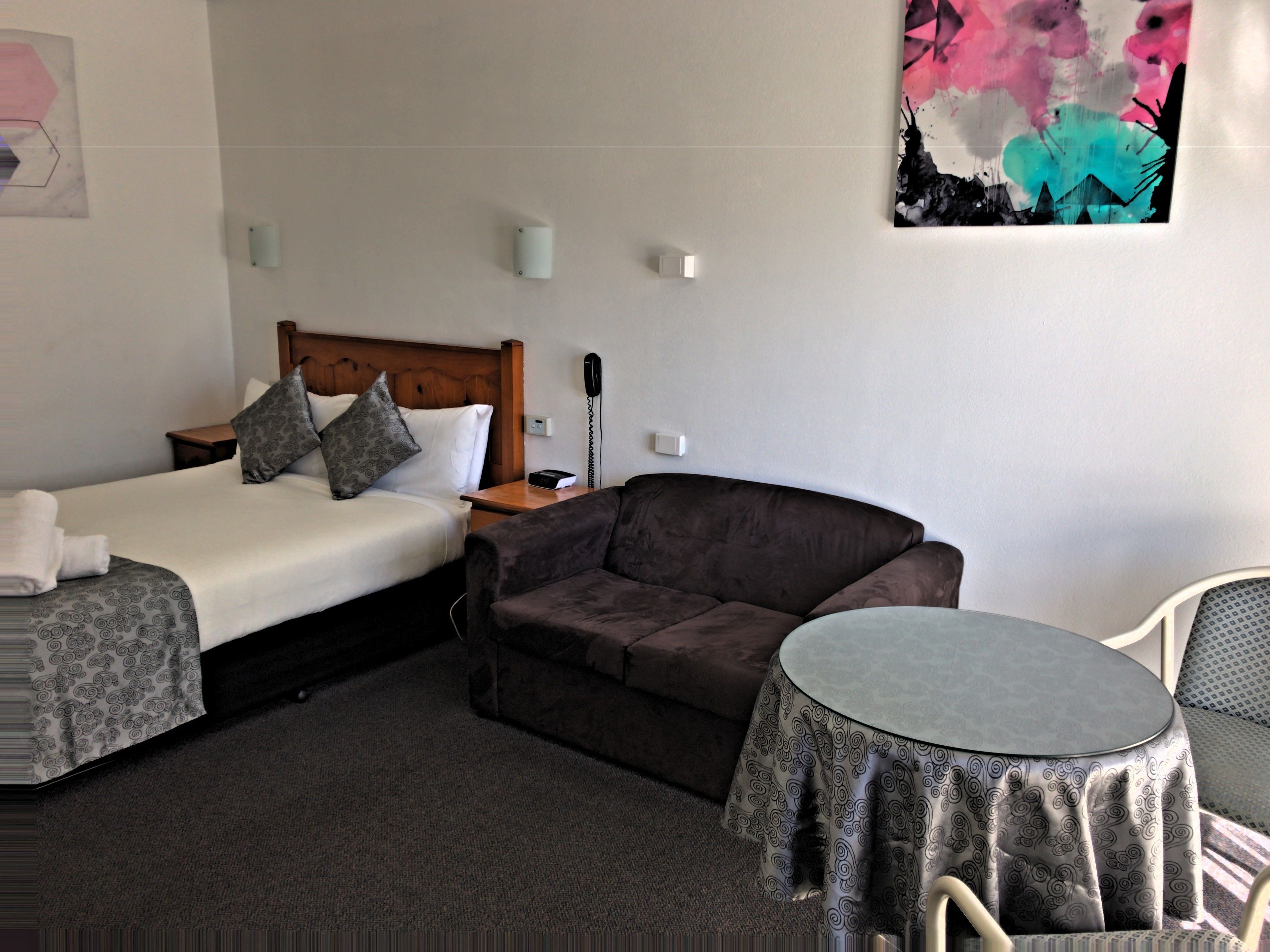 Habitación de hotel con cama, sofá, una mesita y una silla. Hay un teléfono y un enchufe en la pared.