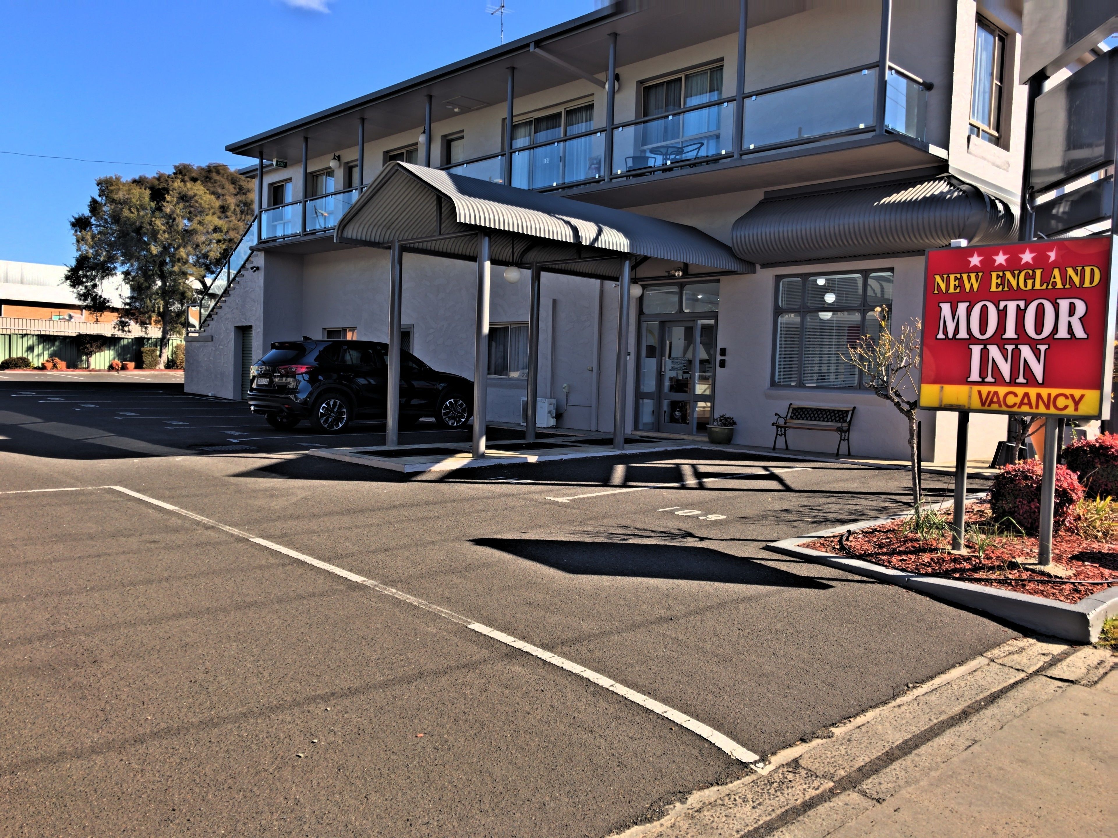A motel with a covered parking area, a signboard advertising a vacancy, and a black SUV parked in front.