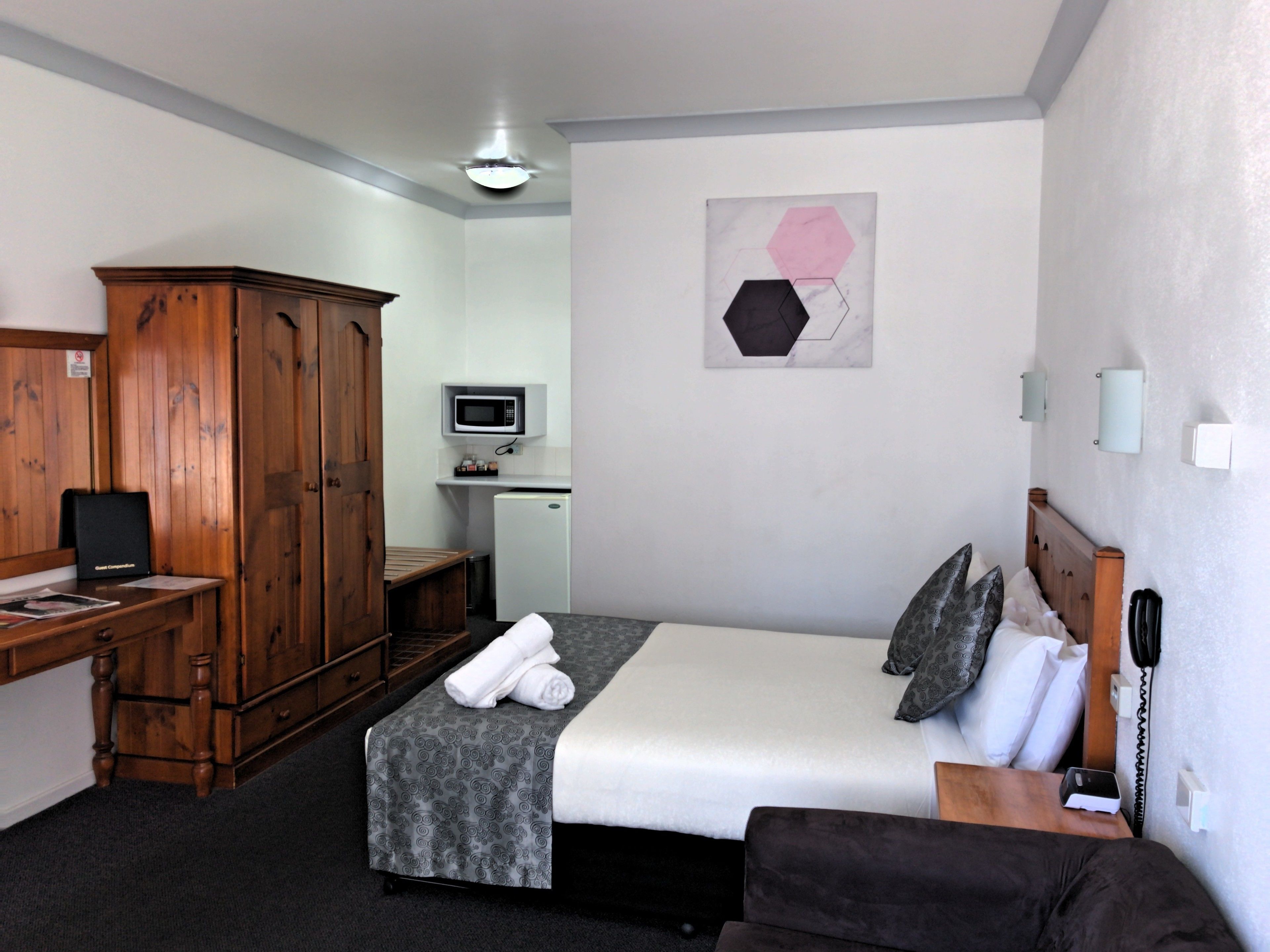 A hotel room with a bed, a desk, a wardrobe, a small kitchenette, and a wall-mounted artwork.