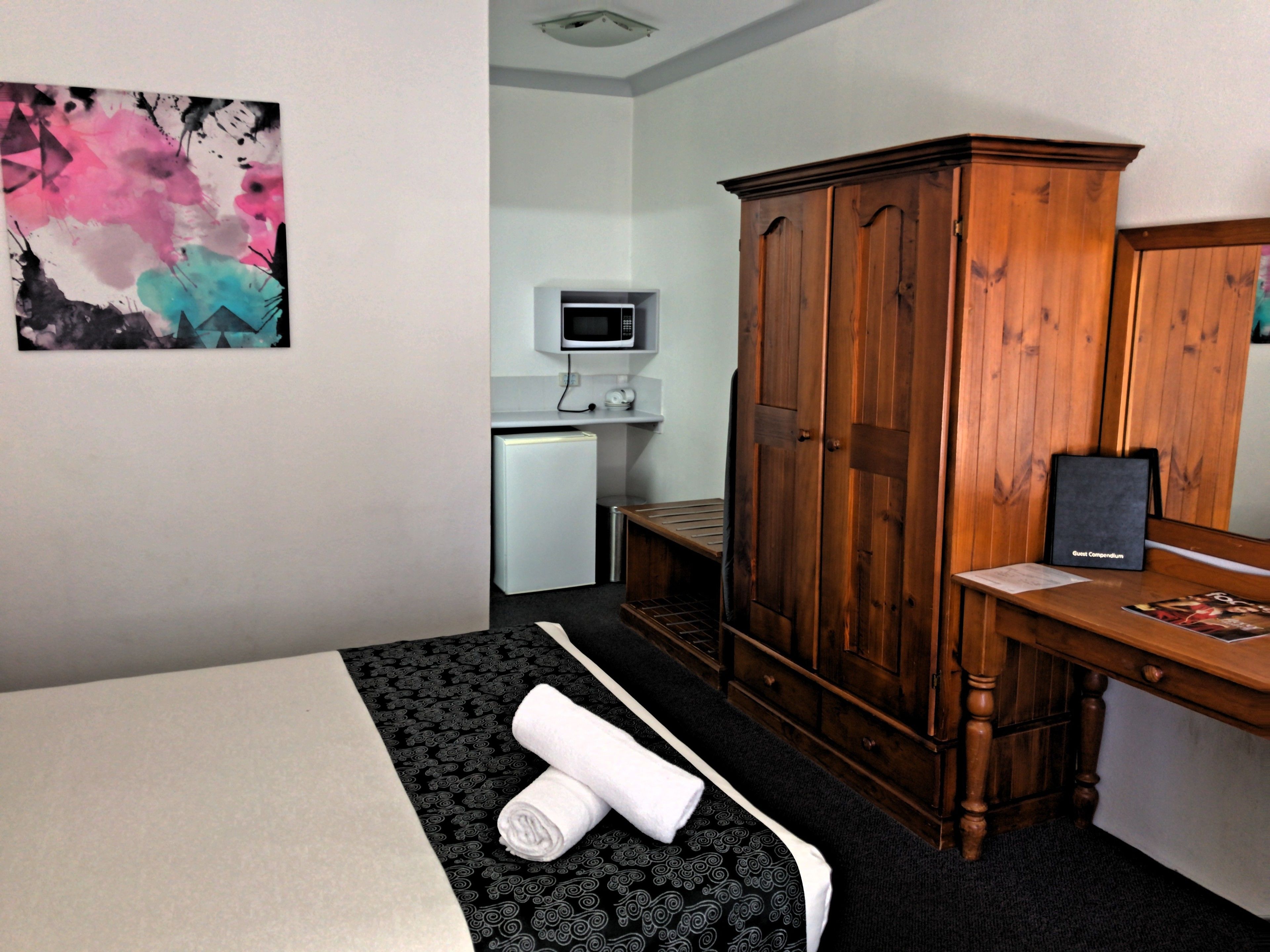 A hotel room with a bed, a wooden wardrobe, a desk with a computer, a small fridge, and a painting on the wall.