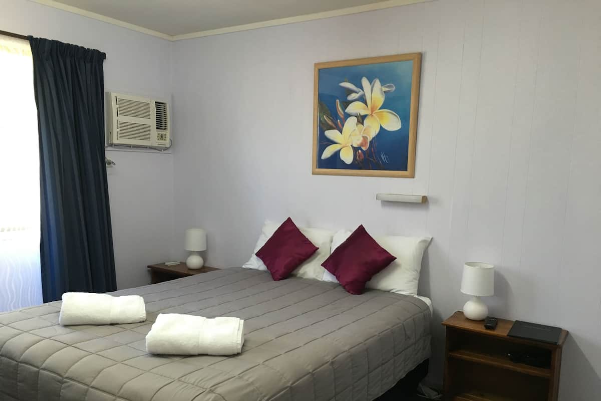 A hotel room with a bed, two nightstands, a painting, and an air conditioning unit.